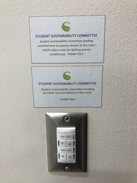 Two signs are displayed above a light switch; Sign 1: "Student Sustainability Committee funding provided new occupancy sensors in this room which reduce costs for lighting and air conditioning. Thank you!"; Sign 2: "Student Sustainability Committee Funding provided new LED lighting in this room. Thank you!"