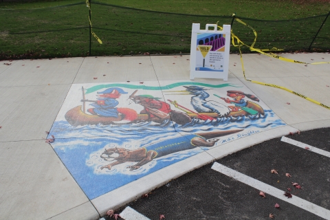 A mural of a a fox (the captain), a beaver, a pelican, and a toad rowing a canoe. An otter swimming under the canoe with a blue onesie that says "Nadiators". There's a sign behind the mural that says "Your Storm drains are becoming more colorful to remind us to put all trash in its place"
