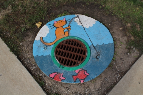 A mural on the circular slab of concrete around a manhole. The mural depicts a cat on top of the manhole fishing where the hook of the fishing rod is one of the loops protruding out of the concrete.