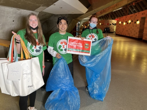 A group of 3 volunteers holding blue recycling bags, signage, and a giveaway tote bag.